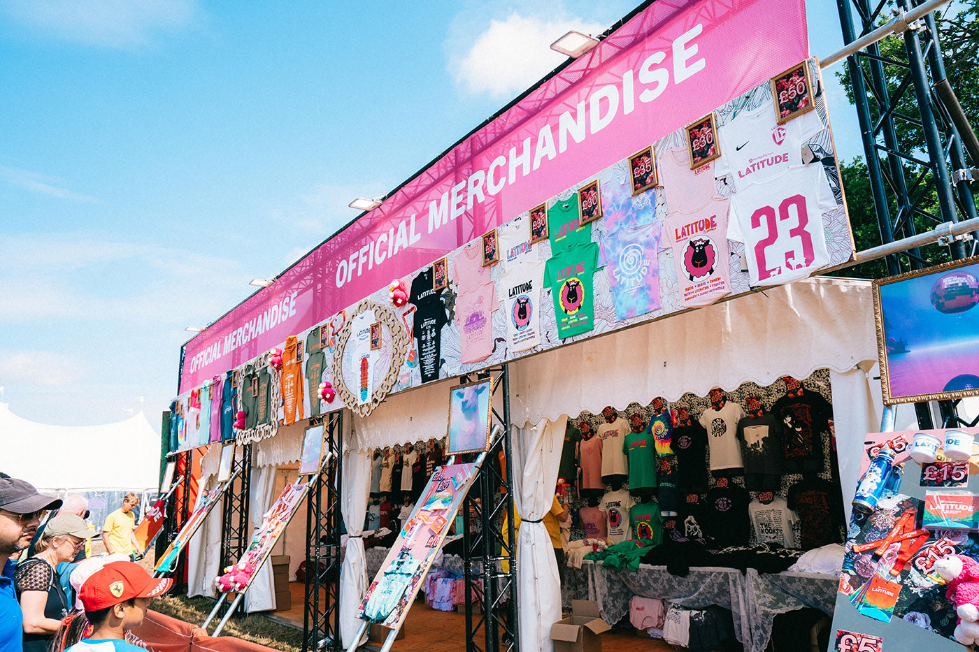 A colorful outdoor tent labeled Official Merchandise displays various shirts, posters, hats, and other items for sale, with people browsing in front of the stall under a bright blue sky.