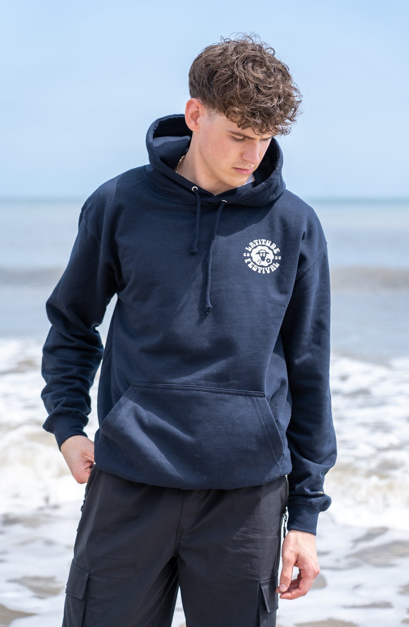 A young woman with curly hair wearing a black hoodie stands by a seaside railing, with beach huts and the shore visible in the background.