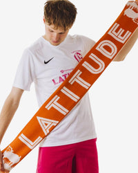 Person wearing a white shirt with a Nike logo and holding an orange 'LATITUDE' scarf.